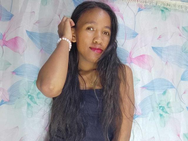 Ashra6_9 stands against a floral-patterned curtain. She has long, straight black hair and wears a black strapless top. Her right hand rests on her head, and she's wearing a white beaded bracelet. Her face is slightly turned to the right, with pink lipstick and a calm expression. The floral curtain features pink and blue leaves with green centers. The image is well-lit, highlighting her smooth brown skin and subtle makeup. The overall composition is simple and centered.