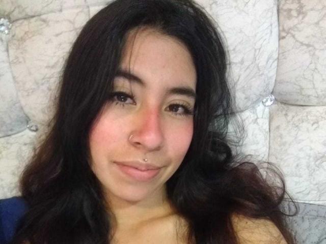 LinaSpearce with dark wavy hair, brown eyes, nose piercing, and lip piercing, smiling softly, sitting on a tufted, white, marble-patterned headboard. Her skin is light brown.
