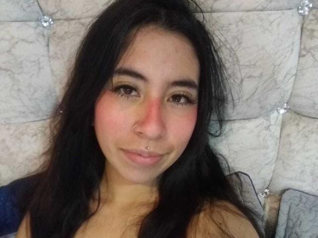 LinaSpearce a de longs cheveux noirs, des yeux marron, un anneau au nez et un léger sourire. Elle porte un eye-liner foncé et un haut bleu, et est allongée contre un tête de lit blanc capitonné..