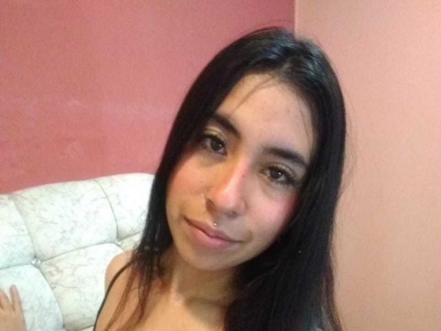 LinaSpearce with dark straight hair, brown eyes, and a small nose piercing, smiling softly at the camera, showing her bare shoulders.