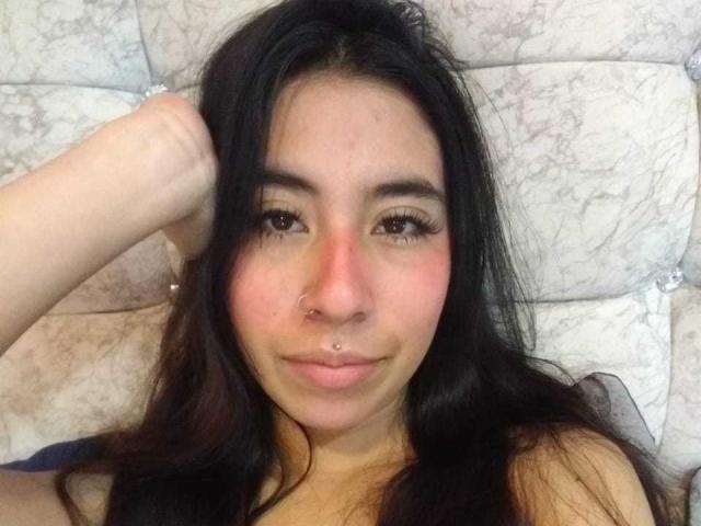 LinaSpearce has dark hair, a nose ring, and a septum piercing, with a hand in her hair, looking at the camera with a slight smile. She's lying on a bed.