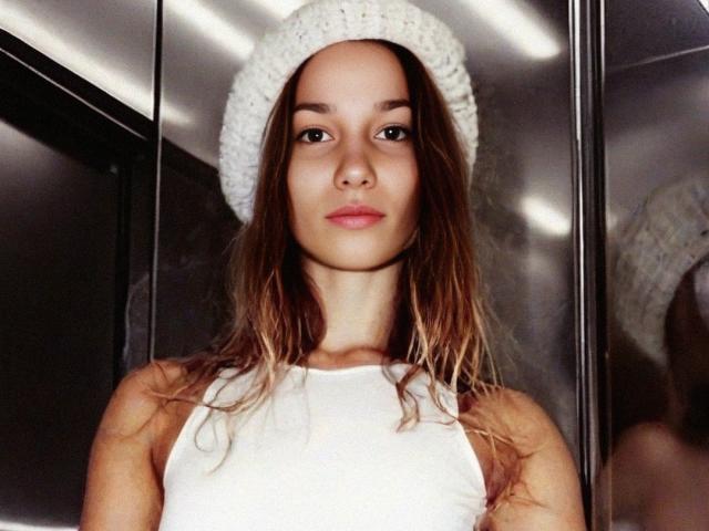 KaiaLein, avec ses cheveux bruns mouillés, porte un bonnet blanc en tricot et un débardeur blanc. Elle a une expression neutre et une légère teinte rose sur les lèvres. Ses seins sont petits, et elle est mignonne..