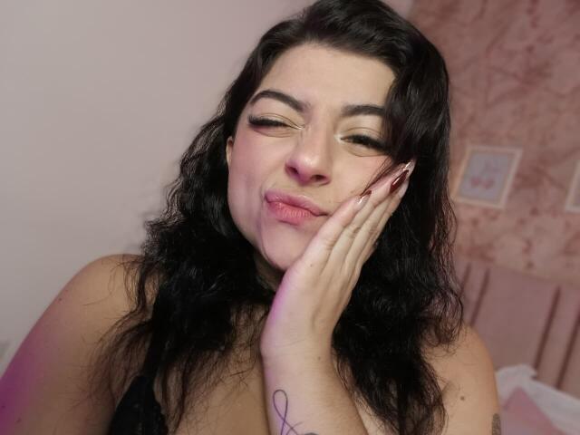 Anntonella Watson, con cabello negro ondulado, ojos cerrados, labios rosados, mano en la mejilla, blusa de encaje negro, tatuaje en la muñeca, una luz rosa tenue y una expresión linda y juguetona..