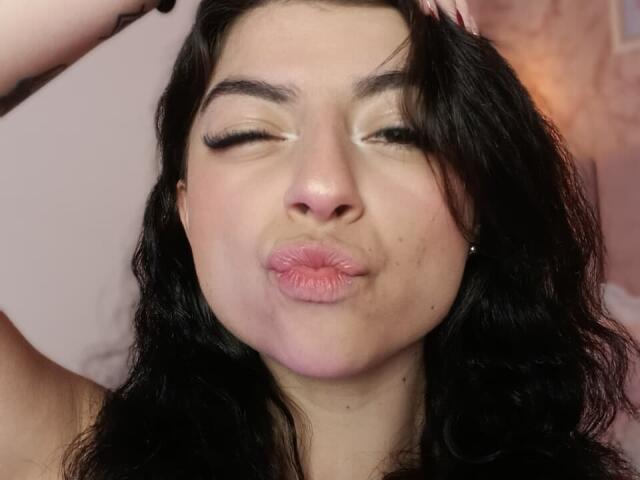 AnntonellaWatson with black wavy hair, eyes closed, puckered pink lips, and hands in hair, looking goofy. She's got a light skin tone and subtle makeup.