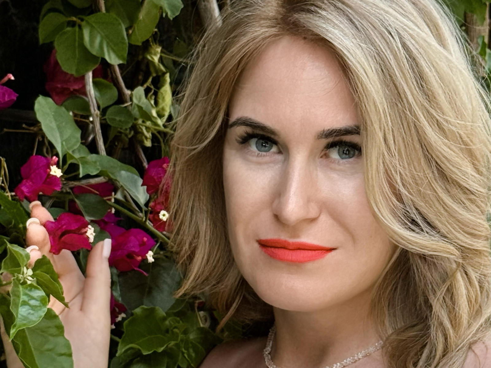 RodieKathlynne with blonde hair, blue eyes, and red lipstick, holding pink bougainvillea, showing a subtle smile, looking directly at the camera. Her tits are not visible.