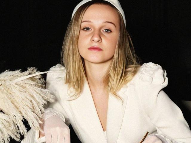 A FlyFreedom tem cabelo loiro, veste um conjunto branco com mangas bufantes, segura uma espanadora branca e fofa, e usa um véu branco na cabeça, olhando diretamente para a câmera..