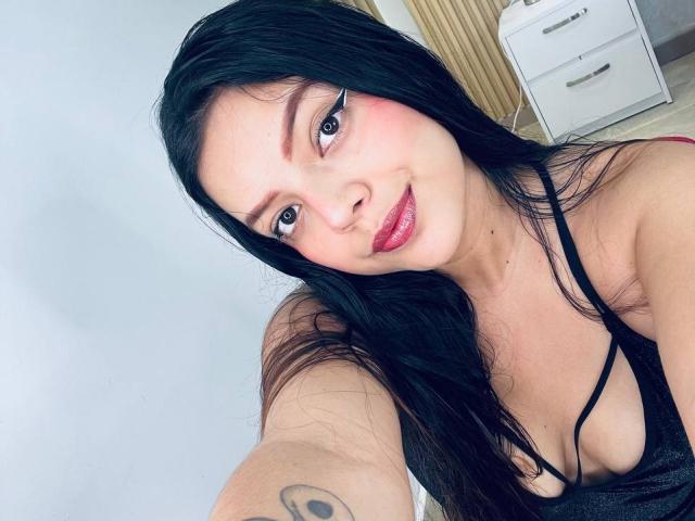ViolettaRouse with long black hair, pink blush, and red lipstick, smiling at the camera, wearing a black spaghetti strap top, showing small tits, tattoo on right arm.