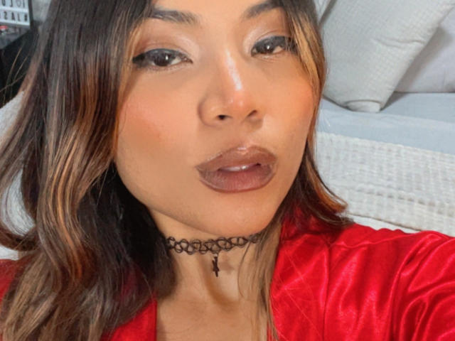 SteffyMiller with brown hair, glossy lips, wearing a red satin top and black choker, looking into camera, slightly pouty face, medium skin tone, subtle makeup.