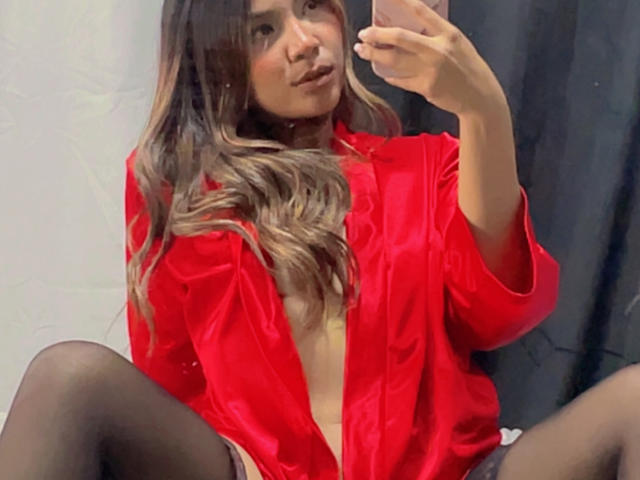 SteffyMiller with long, wavy brown hair, wearing a red shiny shirt, black stockings, and no pants, taking a selfie, showing her bare tits and a small hint of her pussy.