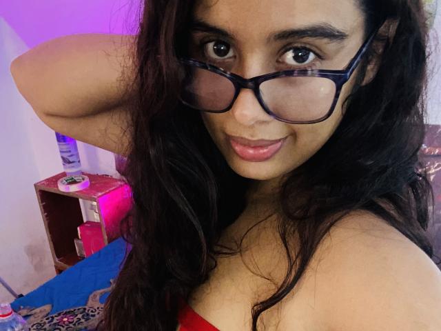 KeyklinOne has brown eyes, long black hair, and wears glasses. She's smiling with pink lips, red top, and her arm behind her head. KeyklinOne's tits are slightly visible.