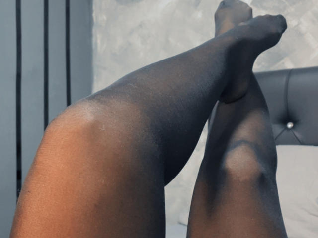 SteffyMiller's legs are crossed, showing her dark brown, sheer black pantyhose-covered thighs and feet, with a subtle sheen on her skin, against a gray tufted headboard.