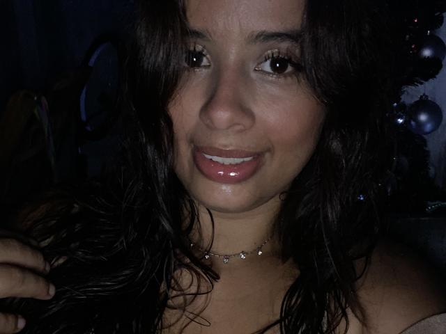 KeyklinOne with glossy pink lips, shiny wet hair, dark eyes, and a subtle smile, wearing a sparkly choker, in dim light, looking at camera with a cute, slightly sweaty face.