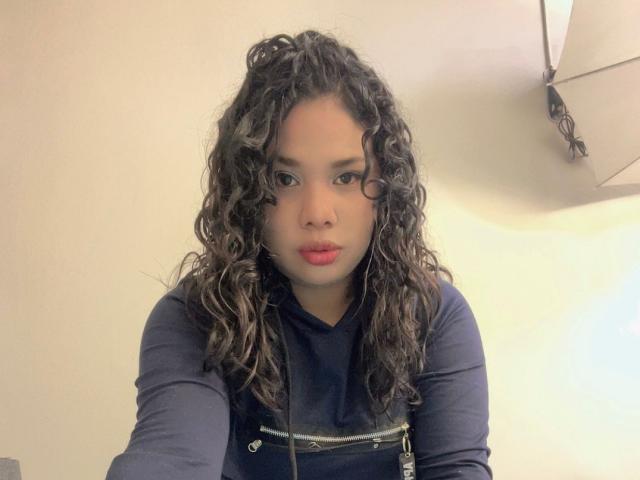 VanessaFory with curly brown hair, wearing a navy blue long-sleeve shirt, has red lipstick, and a serious expression, showing her pouty ass-kissing face in a selfie.
