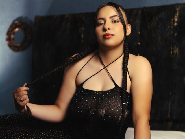 IssabelleHarper with braided black hair, wearing a sheer black dress with criss-cross straps, holding her hair, showing her tits, and red lipstick, in a dimly lit room.