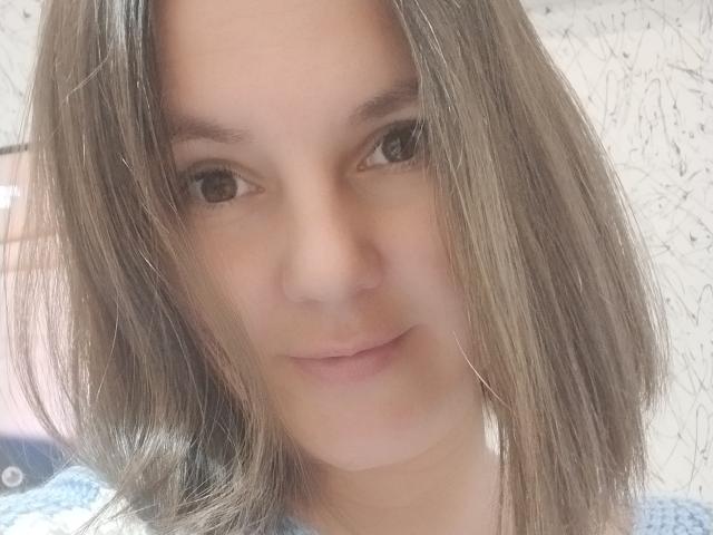 MabelMyriam has straight, medium-brown hair, light skin, and brown eyes with a subtle smile. She's wearing a blue, patterned top, showing her small, natural tits.