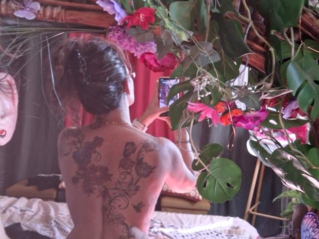 Topless FrenchieLilieFleur with intricate floral tattoos on her back, brown hair in a bun, reaching for a plant with bright pink and purple flowers. Her ass is visible.