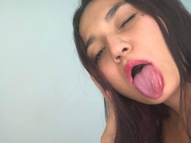 IvyJoness with one eye closed, tongue out, mouth open, showing pink tongue and upper teeth. She has long dark brown hair, thin eyebrows, and a light skin tone.