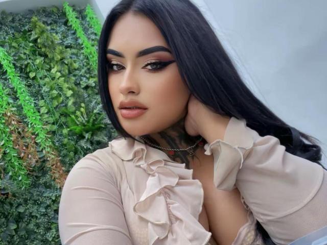 LairaBeamont with long black hair, heavy eyeliner, and pink lipstick, wearing a ruffled beige top, hand on neck, showing tattoos, and a chain necklace, looking seductive.