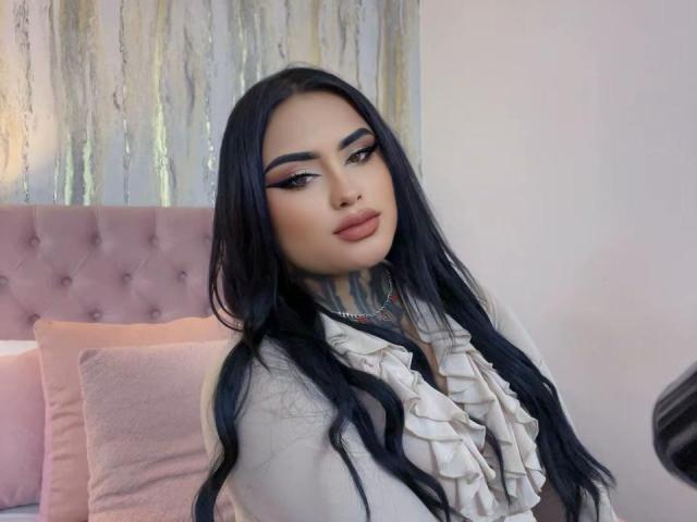 LairaBeamont with long black hair, pink eyeshadow, and red lipstick, wearing a ruffled white top, showing a tattoo on her neck, sitting in a pink bedroom.