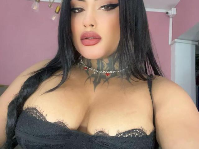 LairaBeamont with black lace bra, showing her big tits, has long black hair, red lipstick, and a spiked tattoo on her neck, wearing a silver chain with red gems.