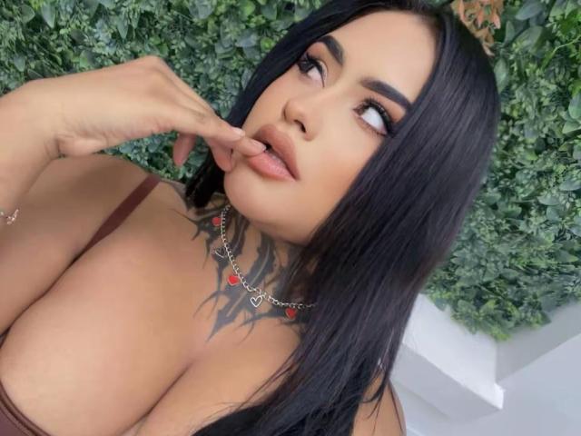 LairaBeamont with black hair, full lips, and a tattoo under her chin, touching her lips with her finger, wearing a red necklace and brown top, showing her large tits.
