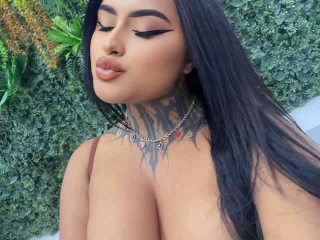 LairaBeamont has long black hair, thick eyebrows, and is puckering her lips with dark eyeliner. She's wearing a necklace with red and heart-shaped pendants, showing her large tits.