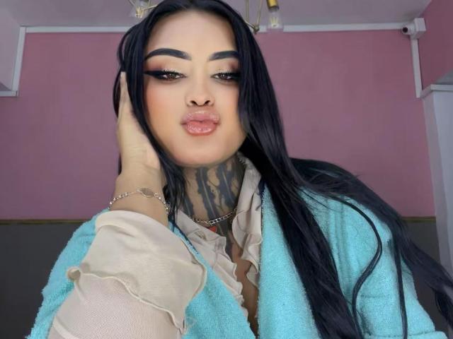 LairaBeamont with long black hair, pouty lips, and thick eyebrows, wearing a blue cardigan, beige shirt, and chain necklace, touching her face, showing a tattoo on her neck.