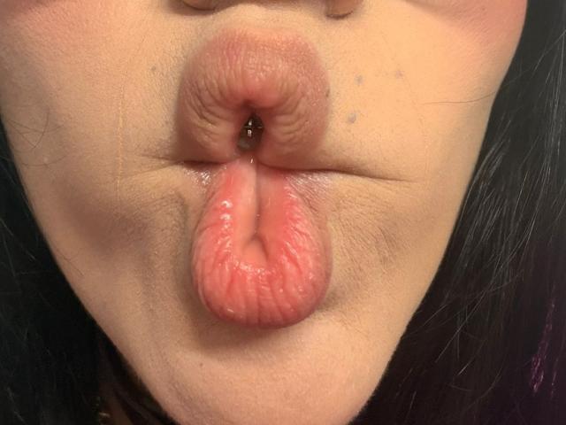 Los labios carnosos, húmedos y rosados de CamilleJuicyHot están fruncidos en un beso, mostrando pequeñas pecas y una textura ligeramente brillante. Su cabello oscuro es parcialmente visible..