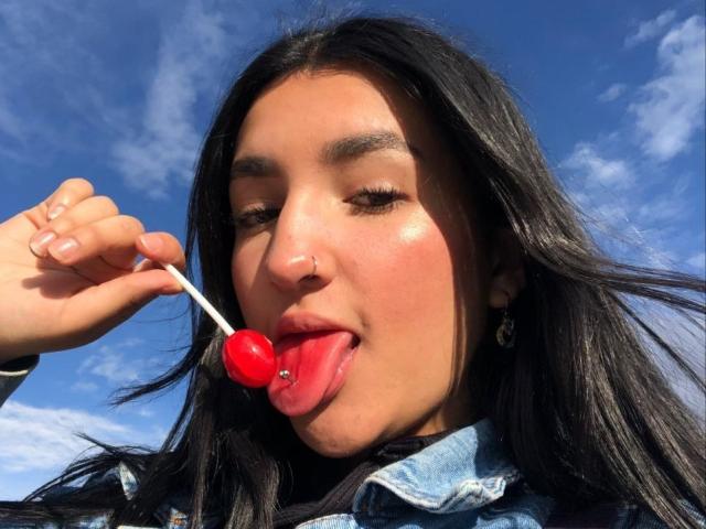 YviTaylor, con cabello negro largo, piercing en la nariz y aretes, lamiendo un caramelo rojo en un palito blanco, fondo de cielo azul, chaqueta de mezclilla y la boca ligeramente abierta..