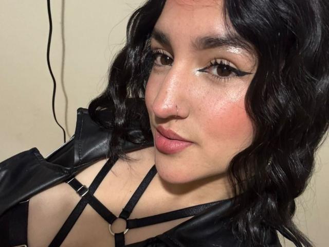 YviTaylor has wavy black hair, black eyeliner, and glossy pink lips. She's wearing a black strappy top with a leather jacket, showing her small tits.