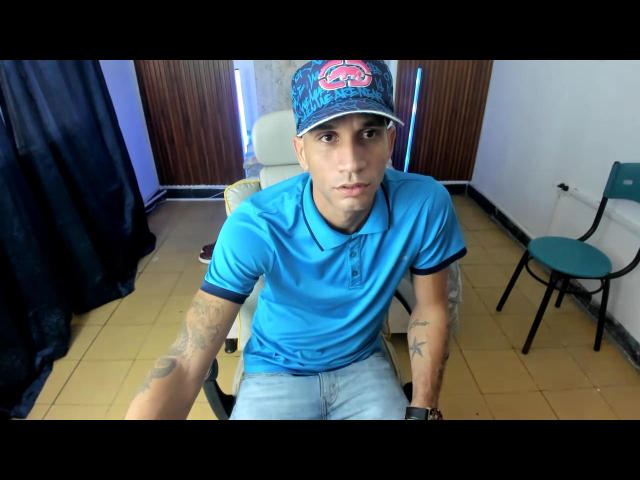 DaniSegura with tattoos on arms, wearing a blue polo, cap with pink logo, and jeans, sitting on chair, looking at camera, yellow-tiled floor, green chair on right.