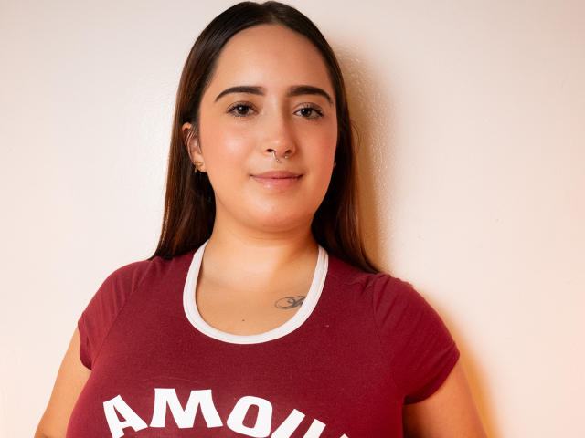 SamySnow, con cabello castaño largo, piel clara, un pequeño aro en la nariz, una sonrisa sutil, vestida con una camisa roja, un tatuaje pequeño en el pecho, de pie frente a una pared blanca, con un pecho ligeramente pequeño, linda, de complexión promedio..