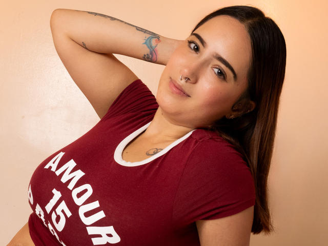 SamySnow, avec ses cheveux bruns, son anneau de nez et ses tatouages, portant une chemise bordeaux, regardant la caméra, les bras derrière la tête, montrant ses seins, un léger sourire, peau claire, en intérieur, pose détendue..