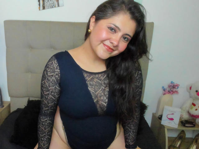 Tiffany, con lunghi capelli neri, indossa una body in pizzo nero, sorride alla fotocamera, mostrando il seno e i glutei, ed è in piedi in una camera da letto con una testiera beige..