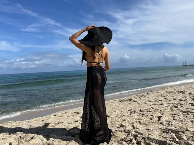A photograph of Bellasweeth standing on a sandy beach with the ocean in the background. She faces away from the camera, wearing a black sunhat, a beige bikini top, and a sheer black dress that reveals her bare buttocks. Her long dark hair is visible. The sky is blue with some white clouds. The water is green with gentle waves. Her right hand holds the sunhat. The sand has a textured, rough appearance. The image uses natural light.