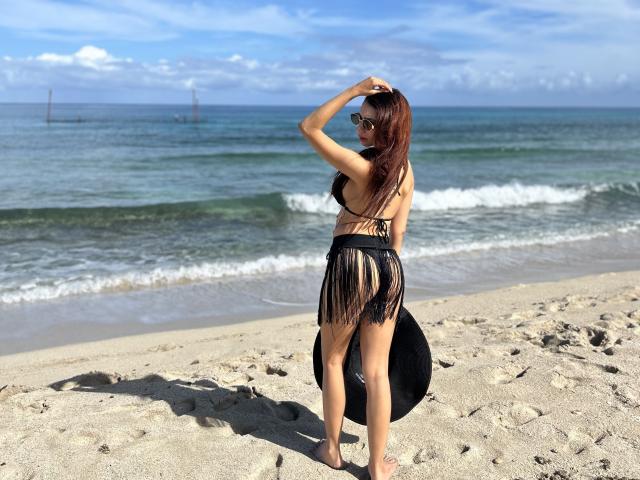 Bellasweeth, com longos cabelos castanhos, vestindo um biquíni preto com franjas, está em pé em uma praia de areia, de frente para o oceano, segurando o cabelo para cima, com o traseiro e os seios à mostra..