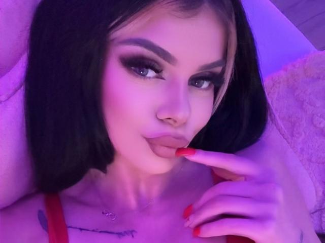 IvyRaven with black bob, heavy makeup, and red nails, pouting with finger on lips, purple lighting, nose ring, and tattoo on chest, wearing red top.