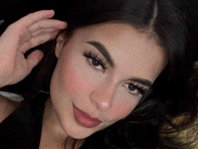 AgataVixen's face with dark brown eyes, long eyelashes, and pink lipstick, slightly smiling, dark brown hair, and fair skin, touching her ear with her right hand.