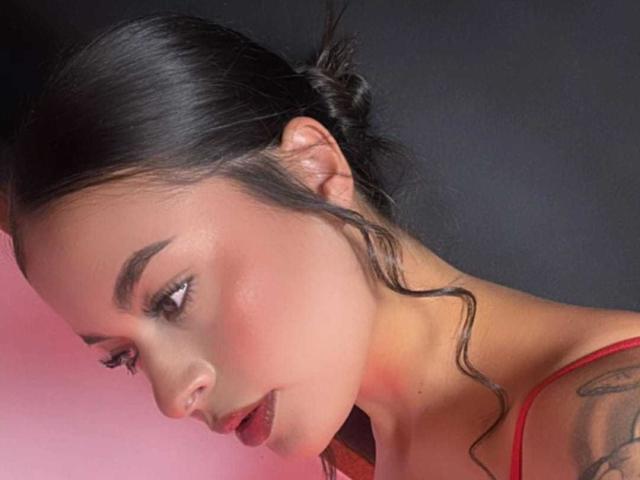 AgataVixen with dark hair in a messy bun, looking down, has red lipstick, a tattoo on her right shoulder, and is wearing a red top. Her face is slightly turned.