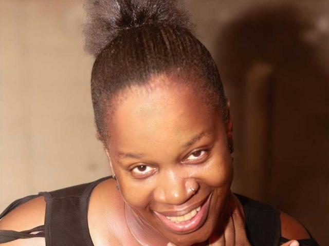 Sexybrexyy con una gran sonrisa, piel oscura y un moño alto. Lleva una blusa negra, se ve linda y feliz, con un ligero giro en la cabeza..