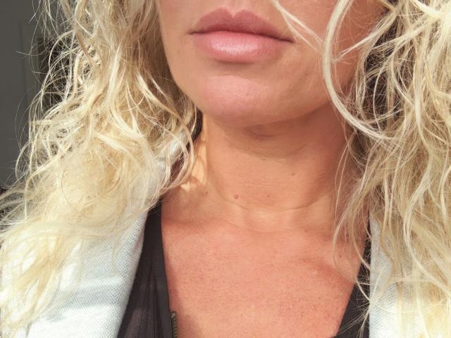 Marie84-hot's face with wavy blonde hair, pinkish lips, and a small mole on her chin, wearing a black and white top, showing her neck and chest.