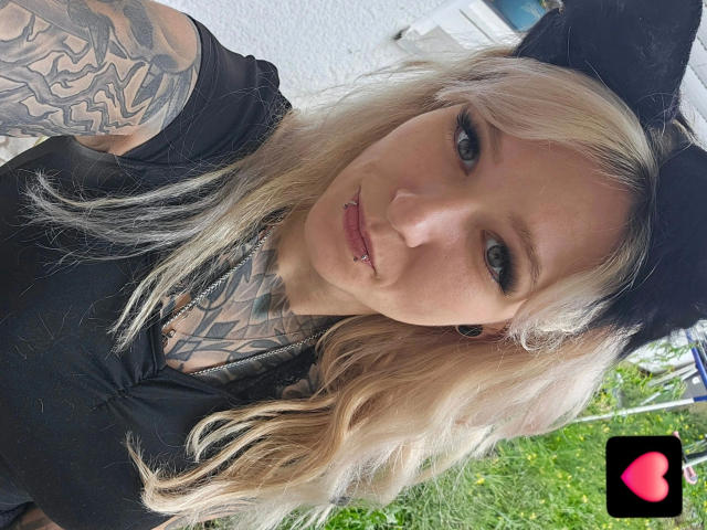 Chrissy-cupcakes-hot with blonde hair, black shirt, tattoos, nose ring, and black beret, showing off her pierced lips and tattoos, standing outside with green grass. Watermark heart icon.