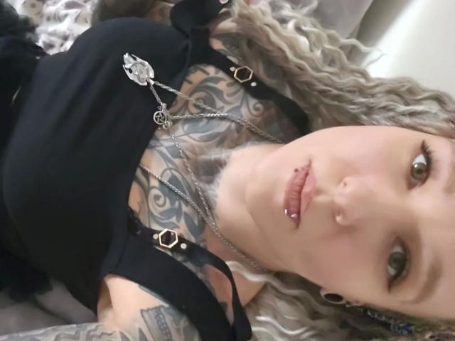 Chrissy-cupcakes-hot has tattoos on her chest and arms, pierced nose, and pierced lips, wearing a black top, with long curly blonde hair, looking at the camera.