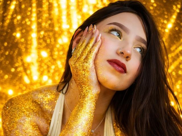 Natasha Vasper met glinsterende gouden armen en handen, donkerrode lipstick, een neusring en lang bruin haar, die omhoog kijkt, haar gezicht aanraakt, op een foto in booru-stijl met gouden, sprankelende elementen..