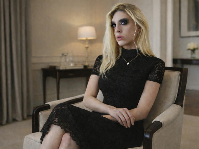 ScharletTsX est une jeune femme blonde avec du maquillage bleu pour les yeux, vêtue d'une robe noire en dentelle, assise dans un fauteuil, avec une expression sérieuse, les mains posées sur ses genoux, dans une pièce..