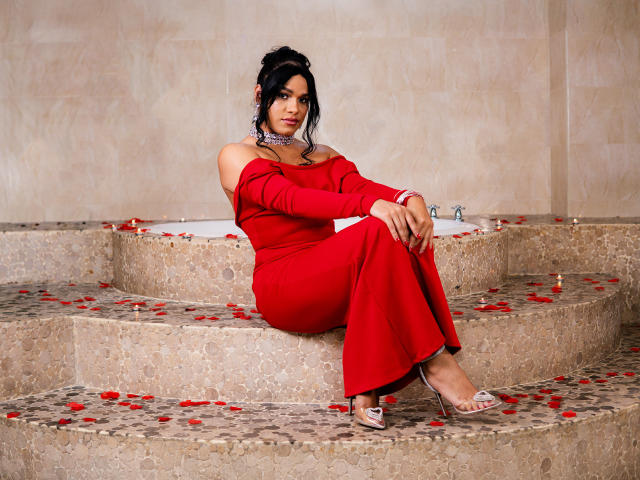 AngelicaRyvera sits on stone steps in a bathroom, wearing a red off-shoulder dress that covers her arms and legs. She has dark hair in a loose updo, wearing large silver earrings. Her right arm rests on her knee, while her left arm is folded. She wears clear high-heels and a red bracelet. Red rose petals are scattered on the stone steps and around the white bathtub behind her. The stone texture is visible, and the background is a beige, tiled wall. The image is sharply focused.
