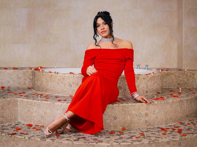 AngelicaRyvera sits on stone steps in a bathroom, wearing a red off-shoulder dress that hugs her curves. Her dark hair is in a loose bun. She has a silver choker necklace and red rose petals scattered around. She's wearing silver strappy heels. Her right arm rests on the steps, while her left hand holds the dress. The background is a beige tiled wall. She looks directly at the camera with a slight smile. The lighting is soft and even.