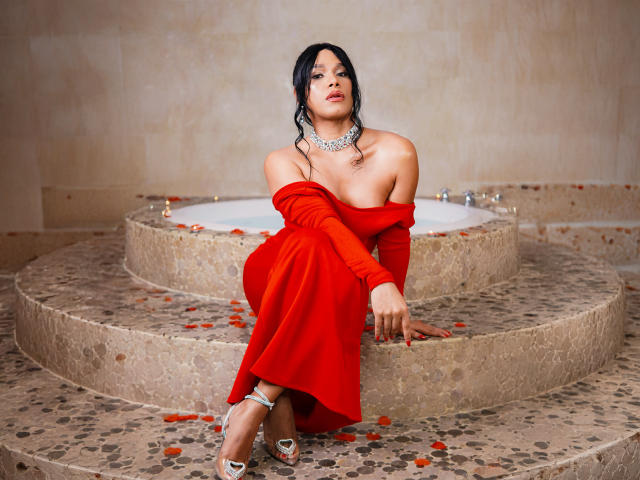 AngelicaRyvera with dark hair, red off-shoulder dress, white heels, and silver necklace, sitting by a circular stone-tiled hot tub, rose petals on floor, looking seductive.
