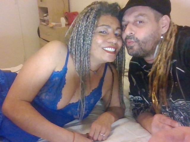 EtnikSatyr with blonde braids and blue lace dress, showing her tits, next to a dude with dreadlocks and black cap, both smiling on a bed.