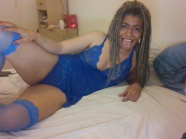 EtnikSatyr with blonde braids, blue lace lingerie, and matching thigh-highs, lying on a bed, tongue out, holding her leg up, showing her ass and small tits.